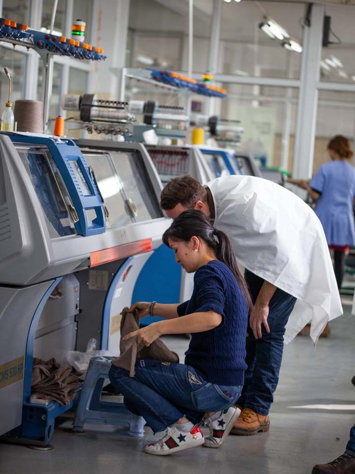 Zwei Personen hocken vor einer Strickmaschine und schauen sich produzierte Wollkleidung an.