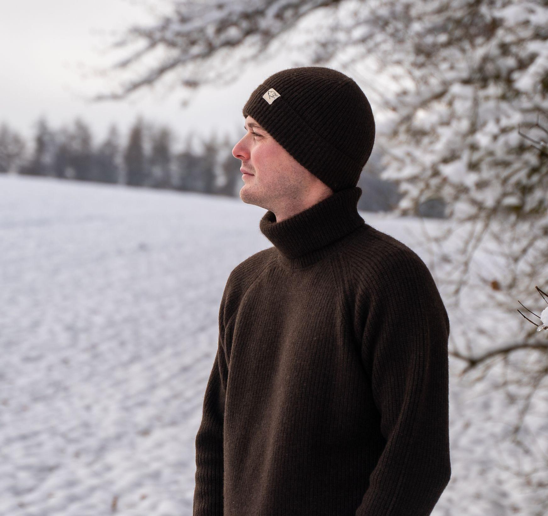 Model steht in Schneelandschaft und trägt dunkelbraunen Rollkragen und Mütze