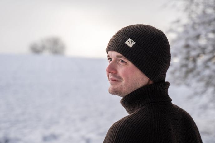 Männliches Modell mit dunkelbrauner Beanie aus Yakwolle in weißer Schneelandschaft.