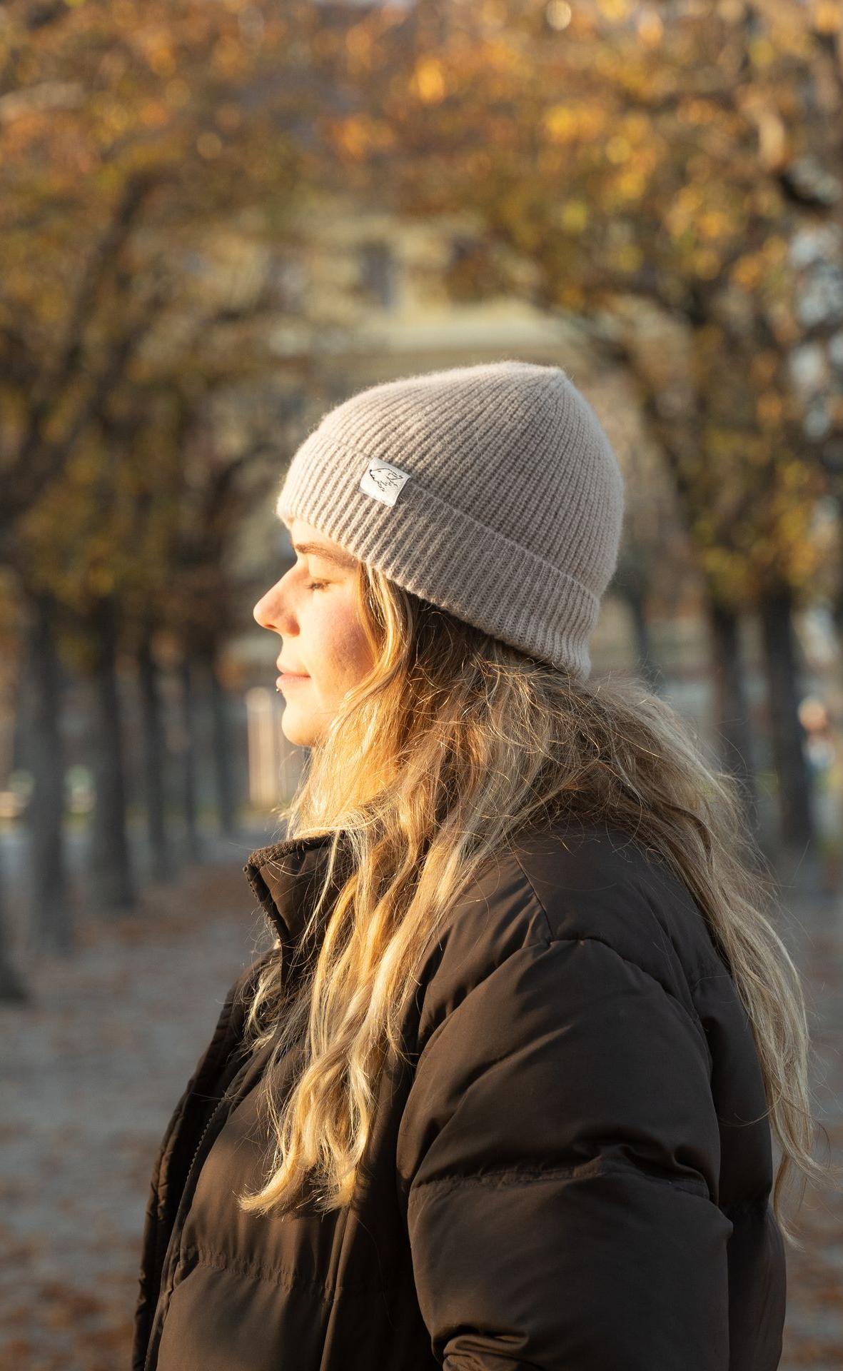 Weibliches Modell trägt beige Beanie aus Kamelwolle und steht in herbstlich gefärbter Landschaft.