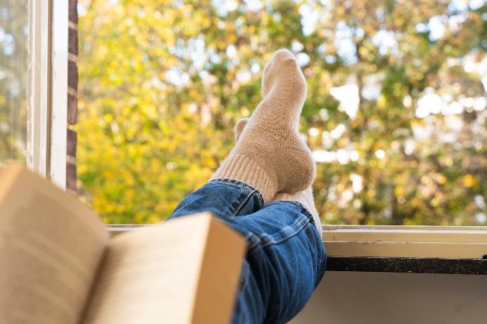 Model trägt beige Socken aus Kamelwolle und liest ein Buch am Fenster.