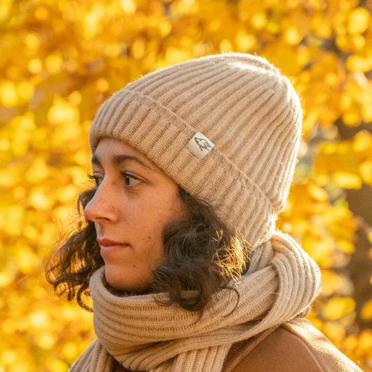 Eine Person mit einer beige gestrickten Mütze und passendem Schal, die nach links schaut, mit Herbstlaub im Hintergrund.