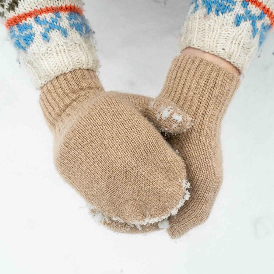 Ein Paar beige gestrickte Fäustlinge mit Schnee auf den Spitzen, getragen von jemandem, der ein weißes Pullover mit blau-rotem Muster trägt.