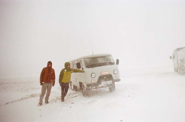 Two people are standing next to a gray car in a snowstorm.