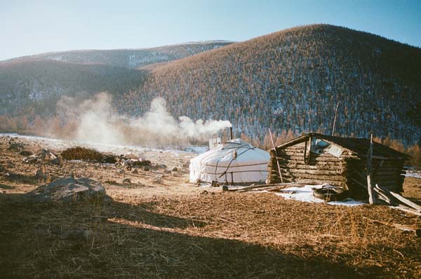 A yurt with smoke stands in the sunlight in front of a mountain