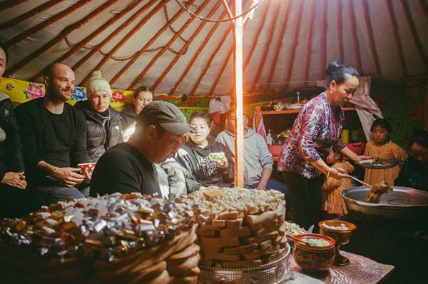 A group of people are sitting in a yurt in front of mountains eating food, a woman is stirring a large pot.