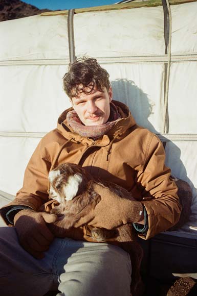 Ein junger Mann hält ein Ziegenbaby im Sonnenlicht vor einer Jurte